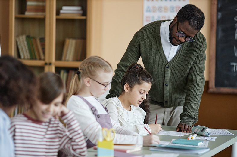 Young African American teacher standing at desk in classroom checking on girl doing grammar exercise during lesson